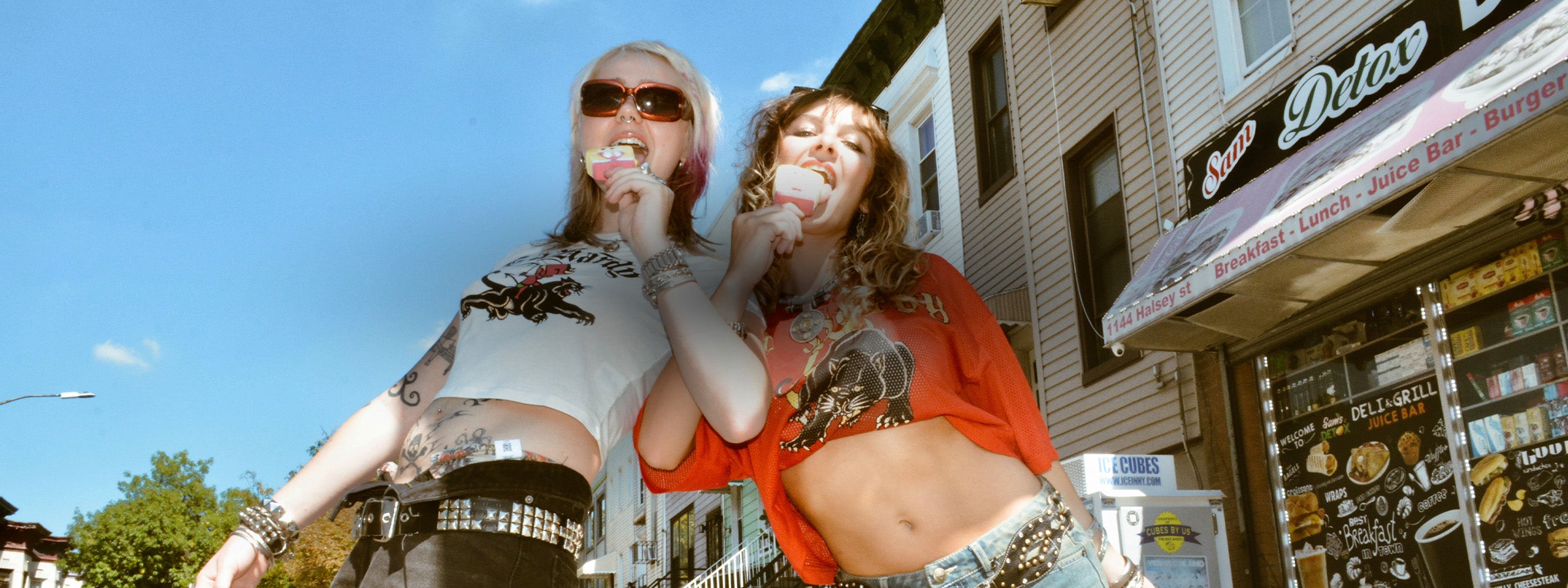 Two women wearing graphic t-shirts standing on a street corner. Both are eating ice cream popsicles, with a building and awning in the background.